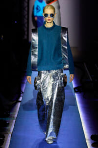 Model on a blue runway wearing a teal high-neck top with metallic silver panels and matching reflective pants, blue sunglasses.