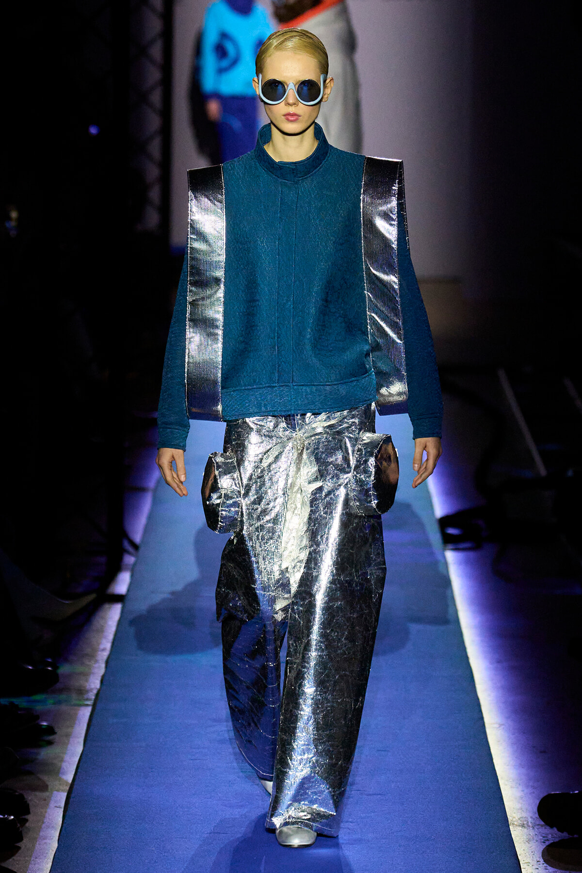 Model on a blue runway wearing a teal high-neck top with metallic silver panels and matching reflective pants, blue sunglasses.