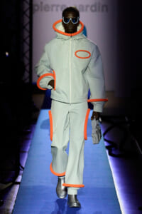 Runway model in pale gray outfit with bright orange piping and circle accents, wearing oversized white sunglasses.