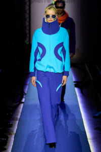 Runway model wearing a bright turquoise jacket with dark blue circular accents and matching pants, oversized sunglasses on a blue-lit catwalk.