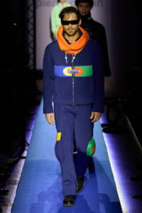 Male model on a runway wearing a navy jumpsuit with a multicolor belt, bright orange scarf, and futuristic sunglasses.
