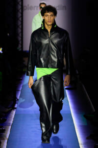 Male runway model wearing a black leather zip-front jacket with neon green collar, paired with matching neon-green pants, walking on a blue-lit runway in a fashion show.