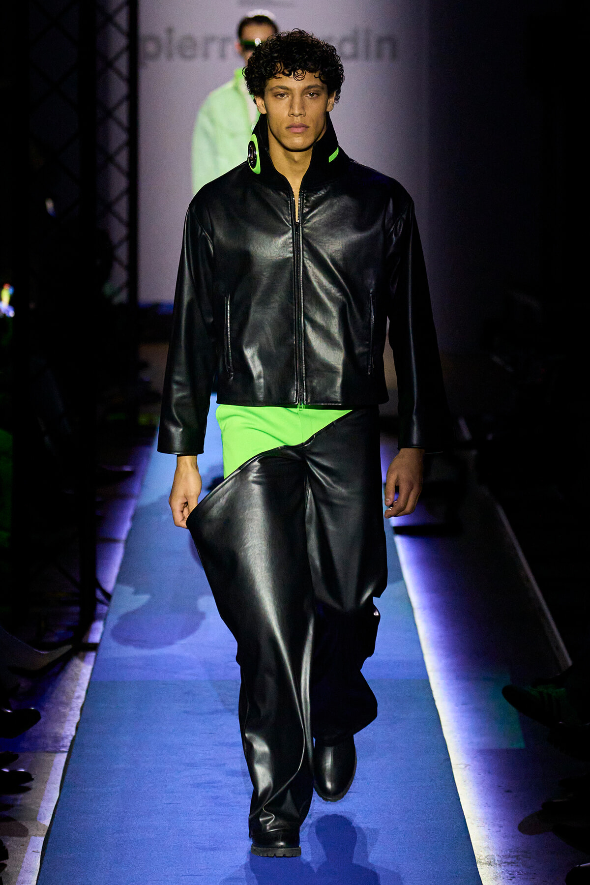 Male runway model wearing a black leather zip-front jacket with neon green collar, paired with matching neon-green pants, walking on a blue-lit runway in a fashion show.