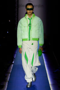 Model walks the runway in a neon green quilted bomber jacket, white pants with green panels, and futuristic green visor sunglasses.