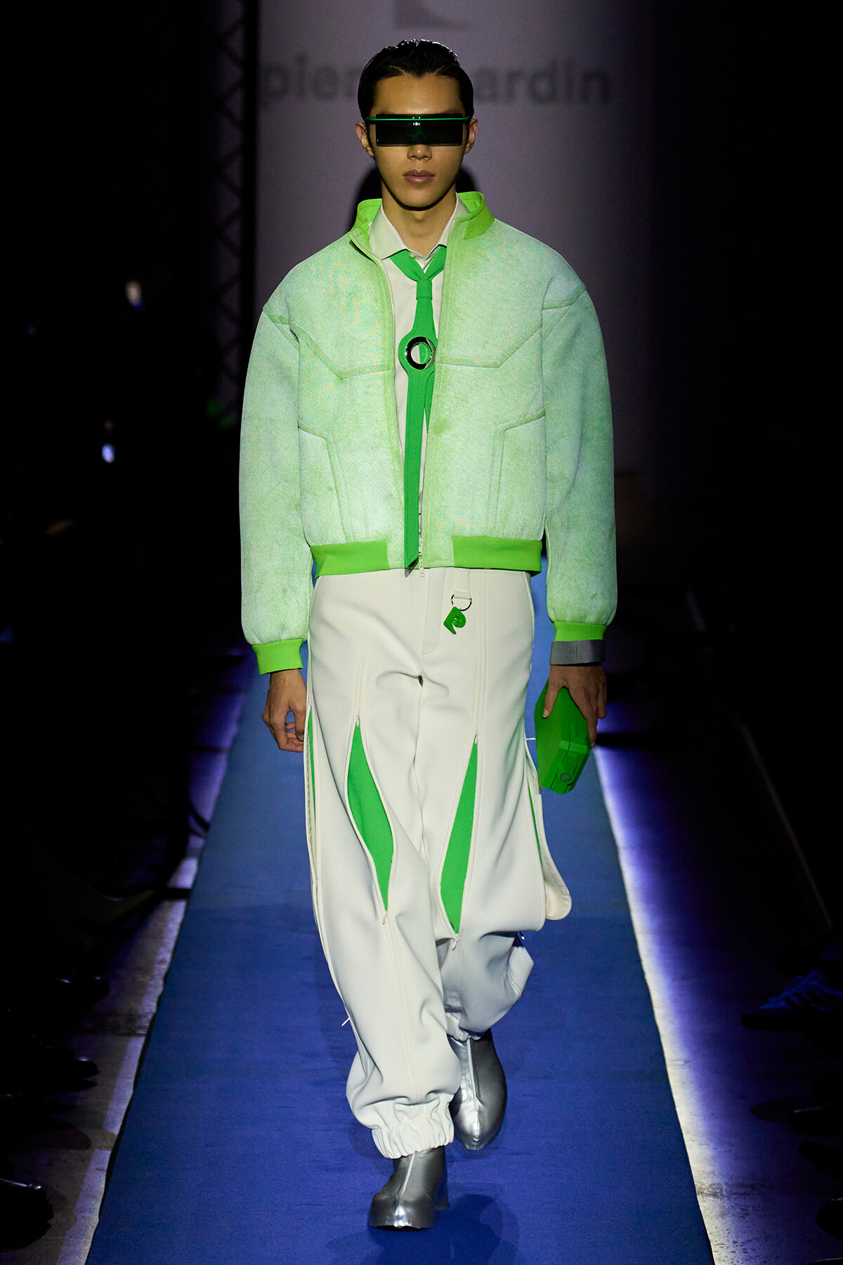 Model walks the runway in a neon green quilted bomber jacket, white pants with green panels, and futuristic green visor sunglasses.