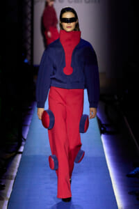 Model walking a fashion show in a blue and red futuristic outfit with oversized collar and circular leg accents, wearing a black visor.