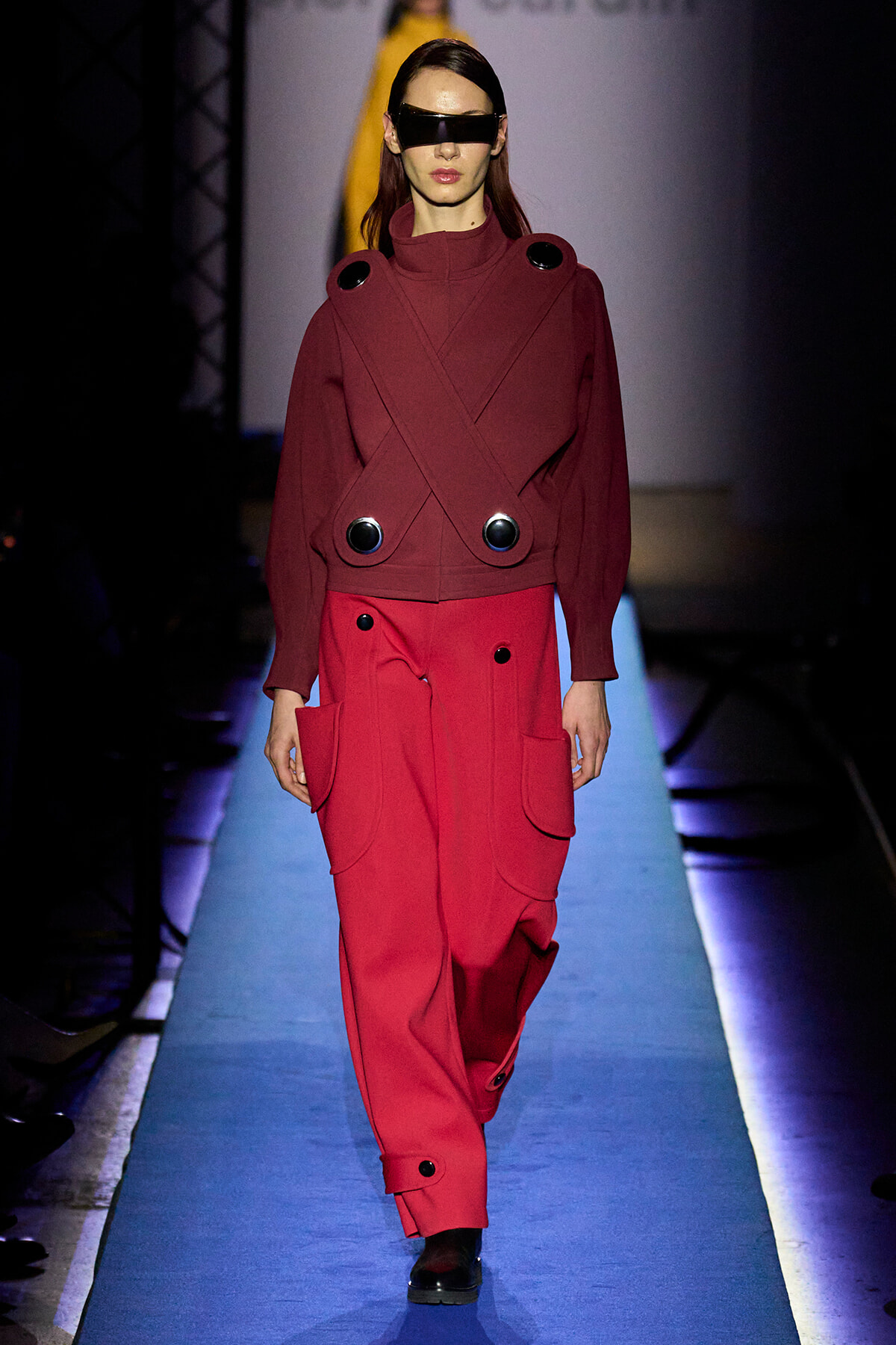 Model on a runway wearing a burgundy wrap-style coat with oversized black circular buttons and red pants, plus a futuristic visor.