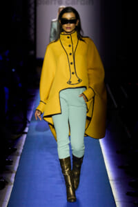 Model on a blue-lit runway wearing a mustard-yellow coat with black piping, light mint pants, and dark visor sunglasses, knee-high boots.