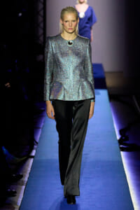 Model on a runway wearing a metallic, crushed-silver top with black trousers on a blue-lit catwalk.