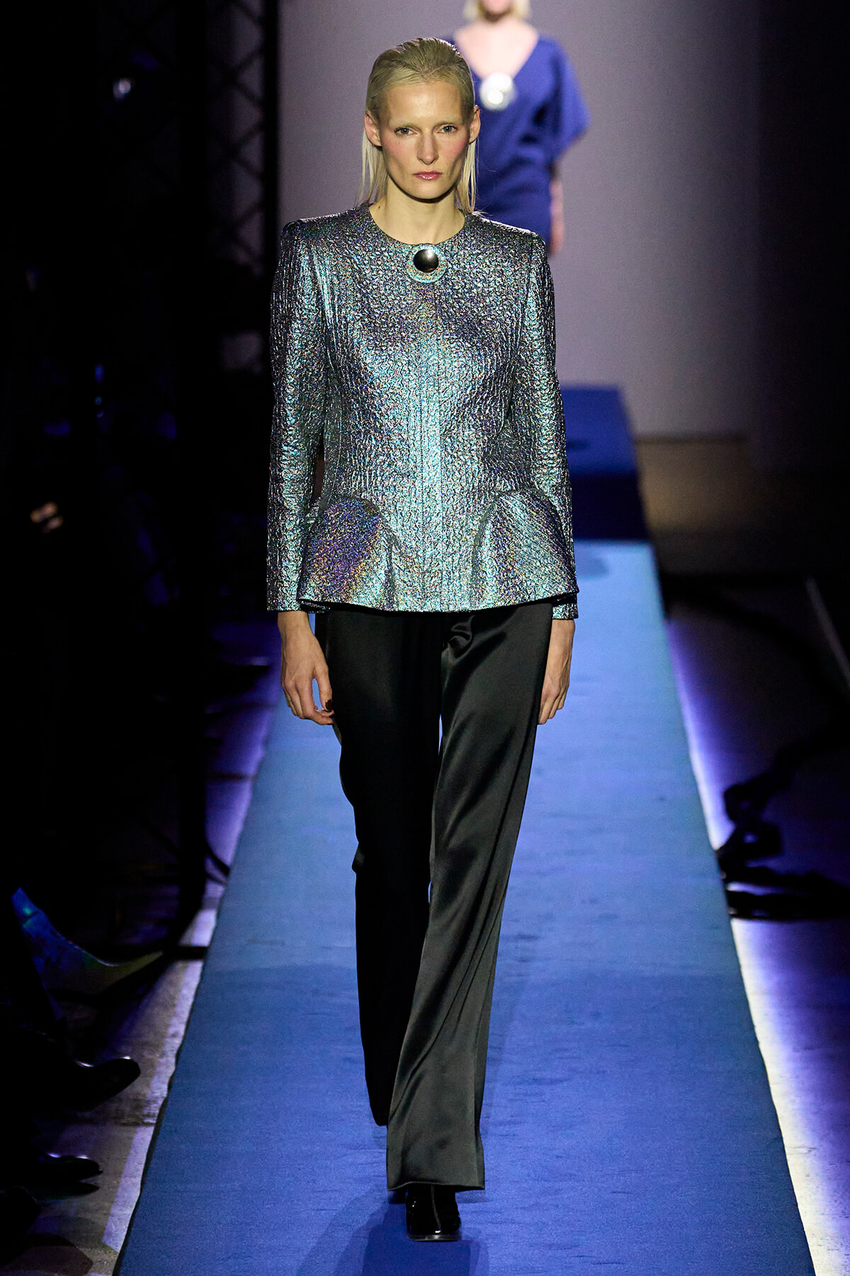 Model on a runway wearing a metallic, crushed-silver top with black trousers on a blue-lit catwalk.