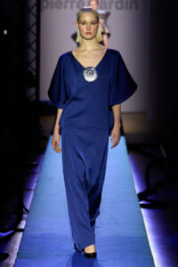 Model walks a blue jumpsuit with a large circular metallic pendant on a fashion runway.