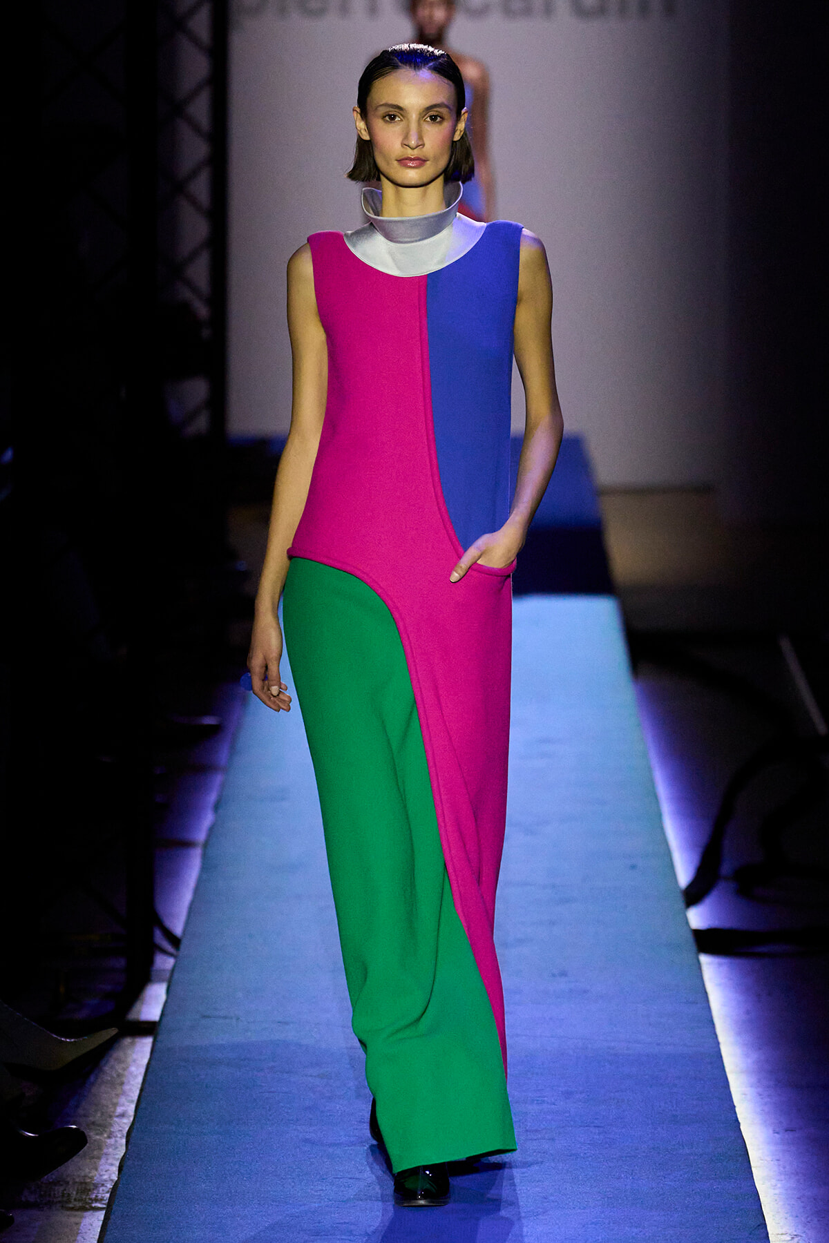 Model walks the runway wearing a color-block sleeveless dress in magenta, blue, and green with a silver collar and a pocket on the right hip.