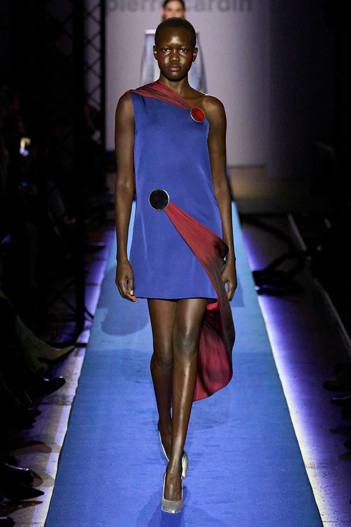 Model walks a blue asymmetrical dress with red sash and circular embellishments on a fashion runway.