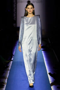 Model walks the runway wearing a light silver satin gown with textured silver panels along the shoulders and sides on a blue catwalk