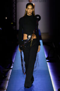 Model walks a fashion runway in a black turtleneck and wide-leg pants, with colorful dangling ribbons wrapped around the torso and shoulder accessory creating a dramatic, avant-garde look.