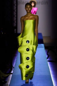 Model walks a neon lime strapless satin gown down a blue-lit runway with oversized black button accents along the skirt.