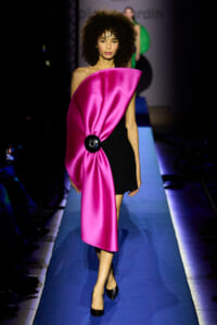 Model on a runway wearing a bright pink satin cape draped over a black dress, fastened with a black circular clasp, walking a blue catwalk.