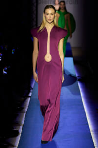Model walking the runway in a magenta floor-length dress with a vertical cutout and circular detail at the waist, navy shoes.