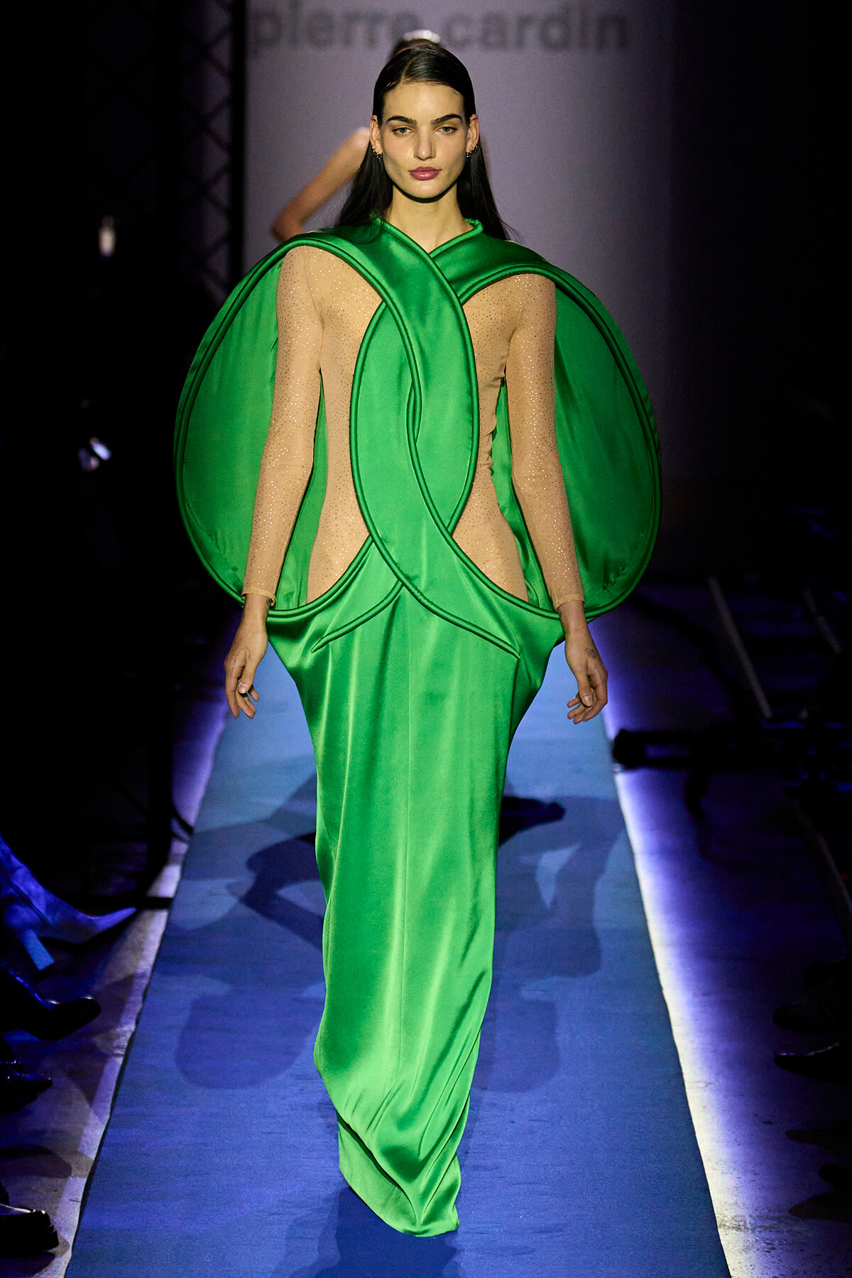 Model walks the runway in a neon-green avant-garde gown with circular sculptural accents on the bodice and shoulders.