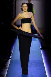 Model walks a fashion runway in a black glitter strapless top and matching glittery skirt, holding a long scarf across midsection.