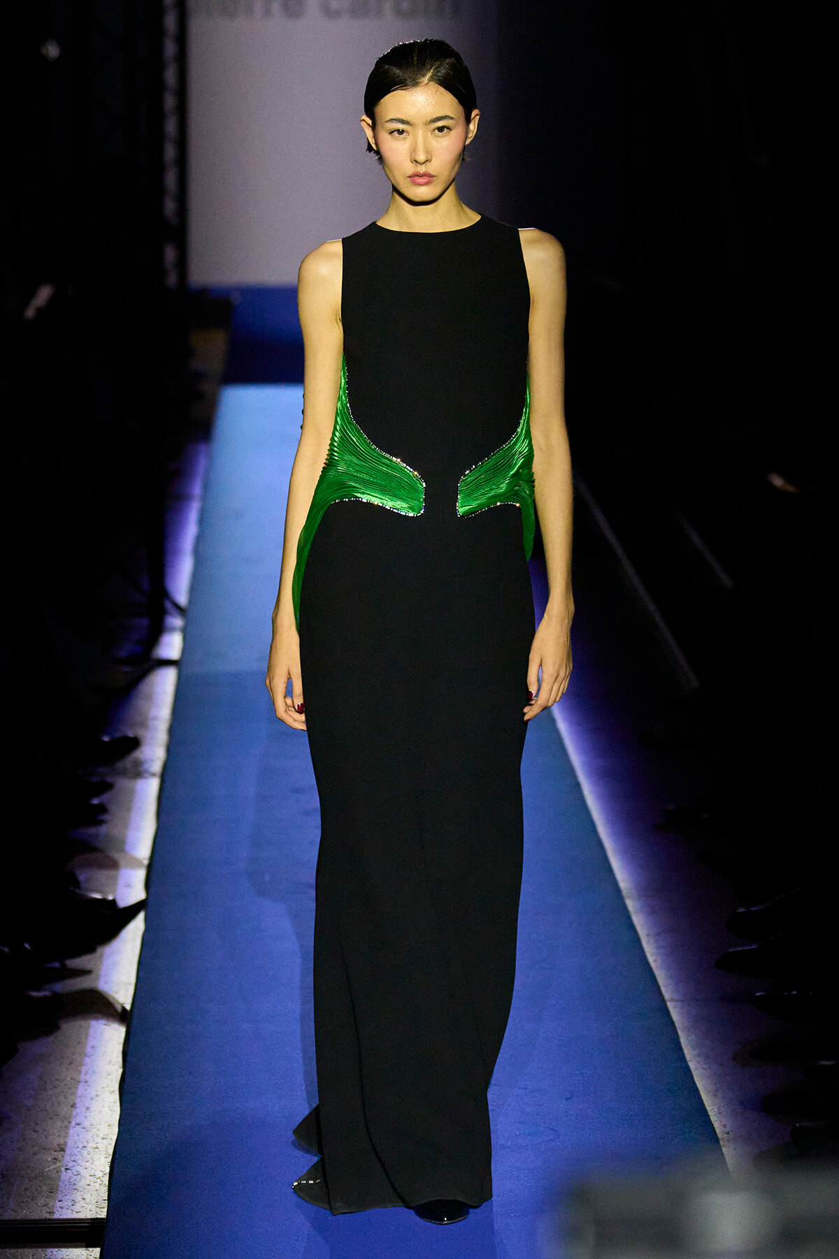 Model walking a blue-lit runway in a long black gown with green metallic side panels on the waist area.