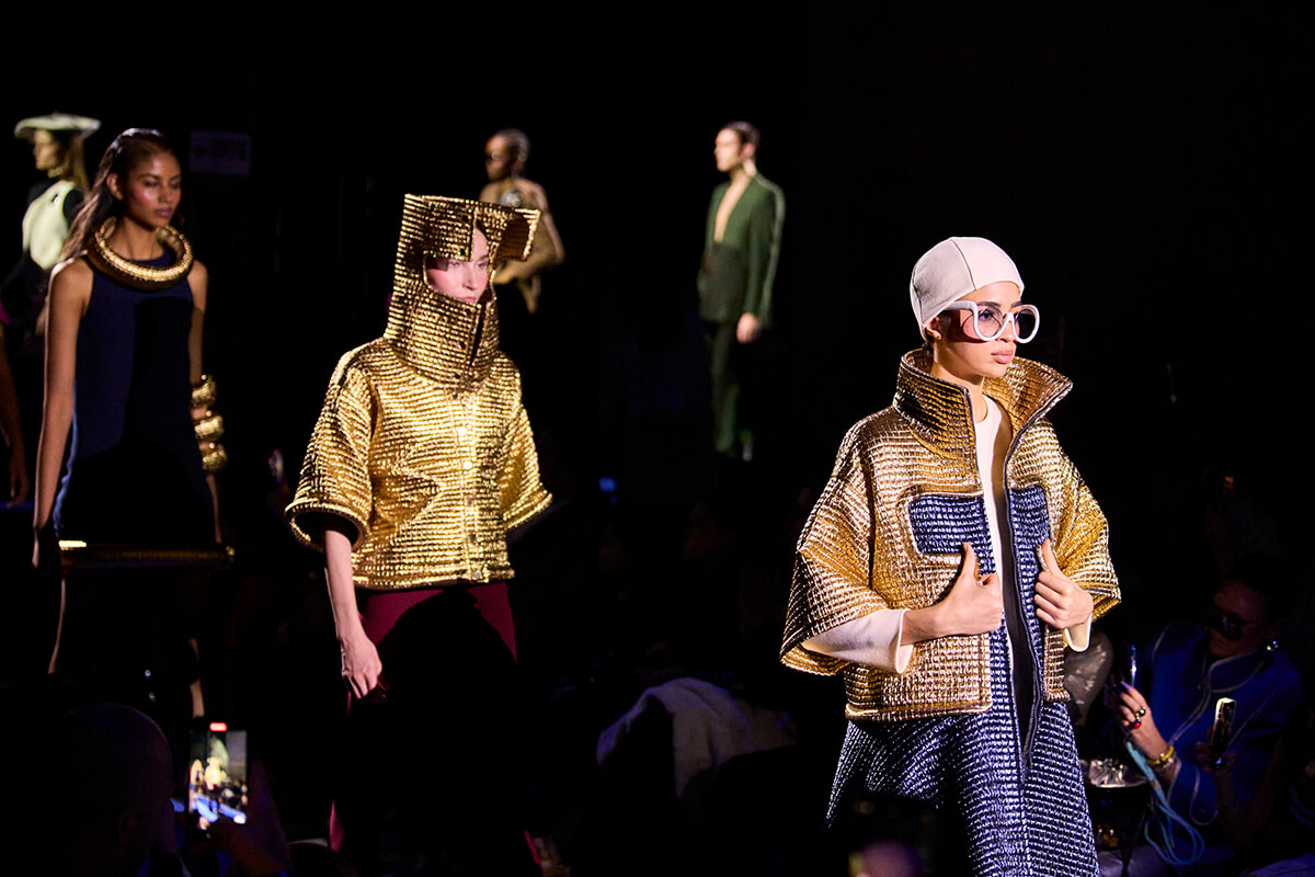 Models walk a fashion runway wearing metallic gold jackets; foreground model wears oversized collar and blue dress beneath, white headpiece and glasses visible on the right