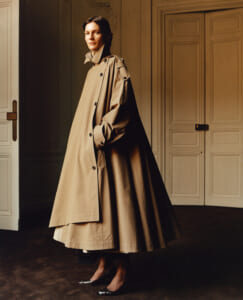 Woman in a long beige trench coat stands in a doorway-lit room, hands in pockets, wearing dark heels.