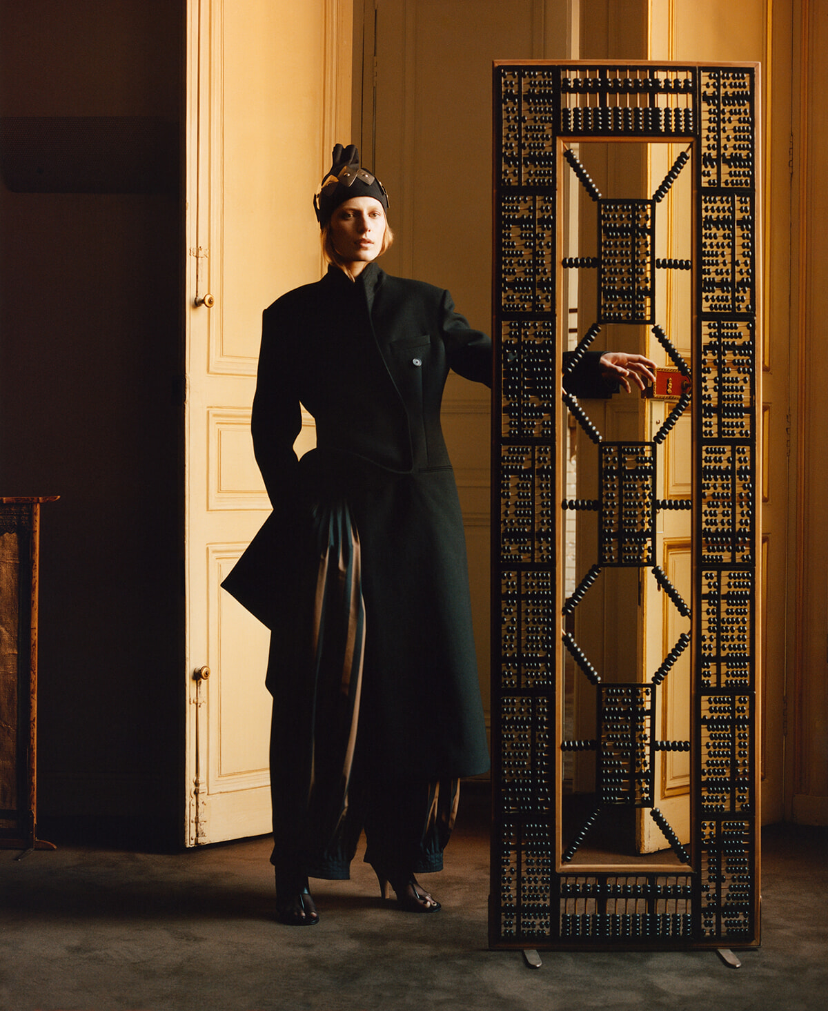 Woman in a dark coat and hat stands beside a tall geometric metal sculpture in a doorway, inside a warmly lit room.
