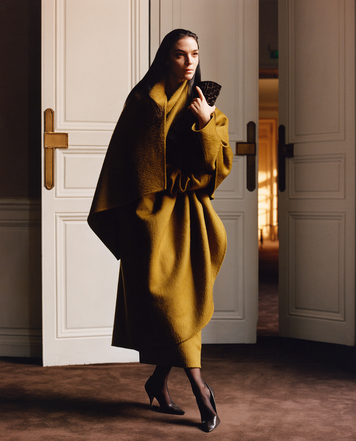 Woman in a mustard coat and black heels entering a room, holding a clutch with a doorway behind her.