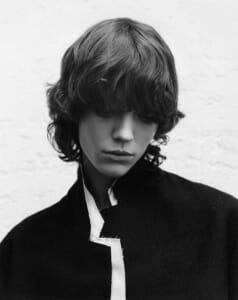 Black-and-white portrait of a young person with shaggy hair looking down, wearing a dark coat with a light trim.