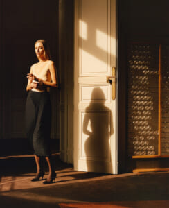 Woman stands in a sunlit doorway wearing a beige sleeveless top, a navy striped skirt, and black heels, with a large shadow on the door behind her.