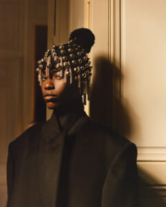 Portrait of a person in a dark coat wearing an ornate beaded headpiece with hanging chains, standing in a warm indoor light.