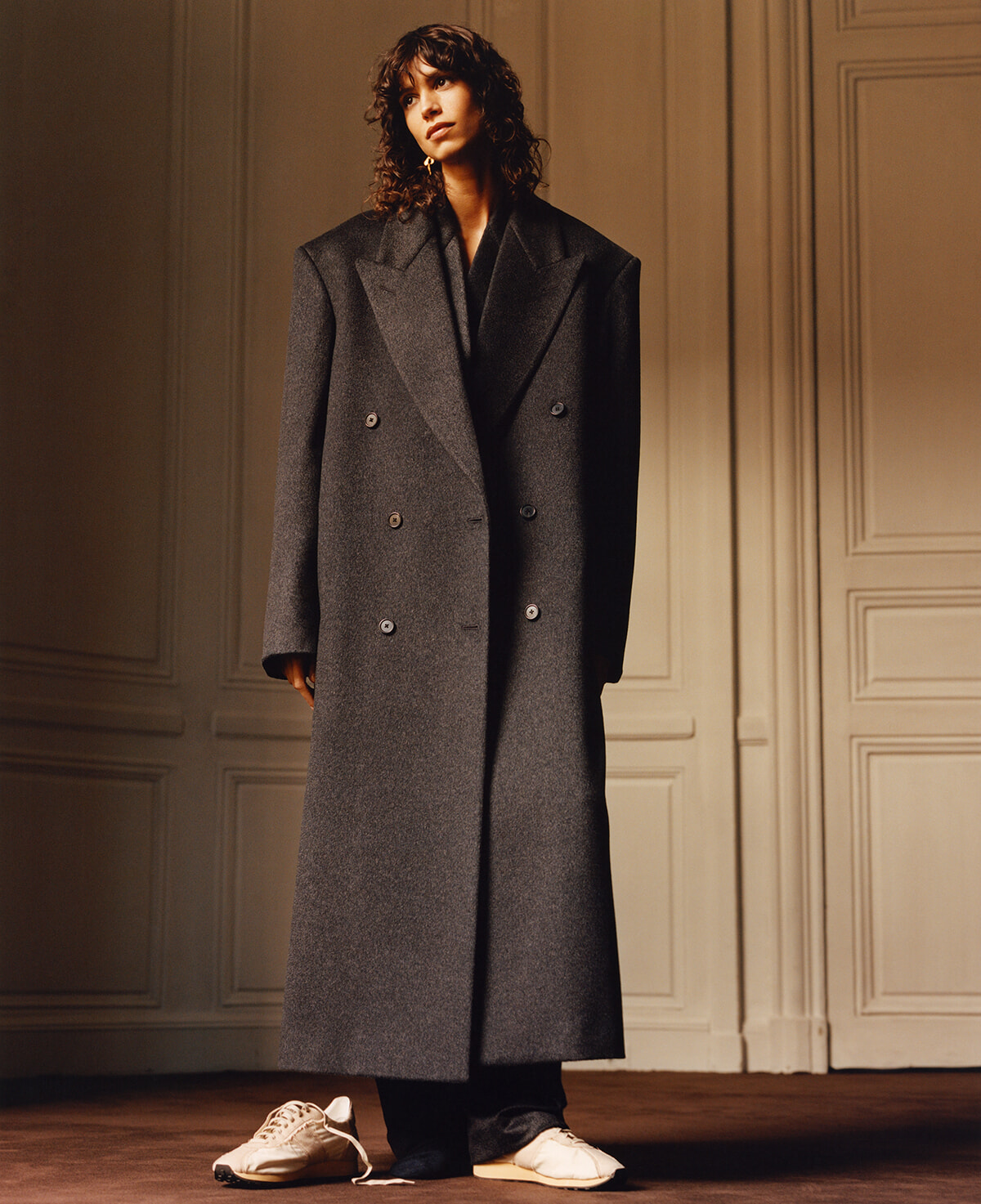 Model wearing a long dark gray double-breasted wool coat with wide shoulders, standing in a warmly lit paneled room, white sneakers visible at the feet.
