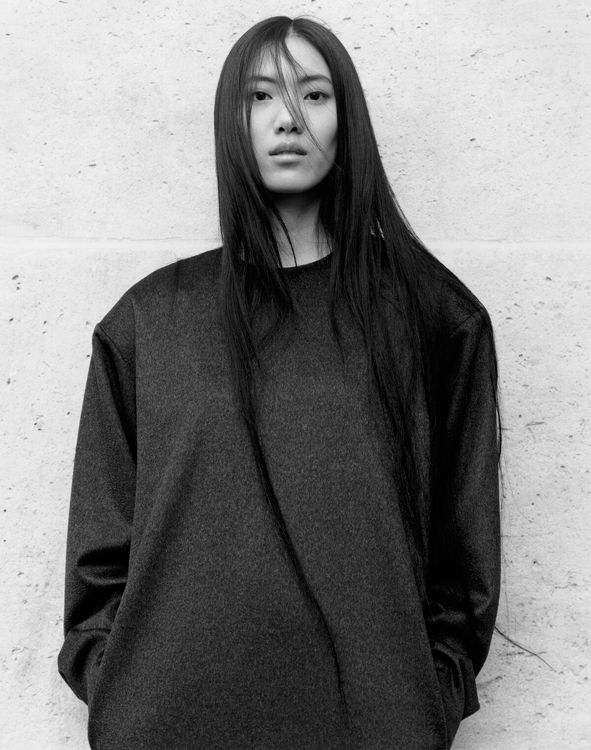 Young woman with long dark hair standing against a concrete wall, wearing an oversized dark sweater (black-and-white photo).
