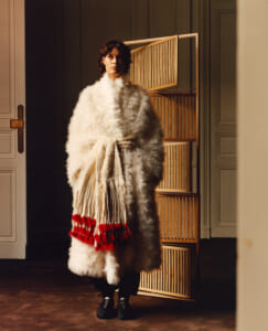 Model in a long white faux fur coat tied at the waist, indoors beside a wooden slatted room divider.