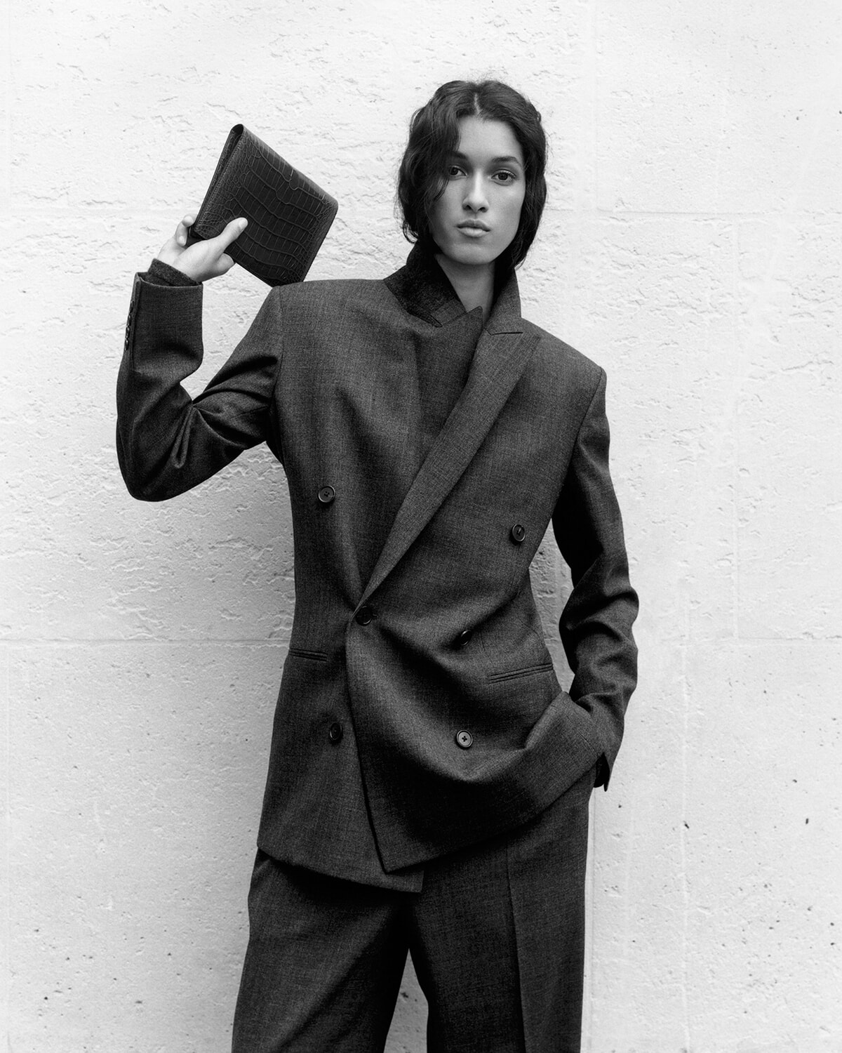 Woman in a tailored double-breasted suit holds a textured clutch aloft against a light brick wall, looking at the camera.