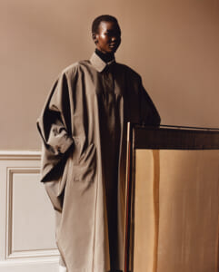 Person in an oversized beige trench coat standing next to a large folding screen in a softly lit room.