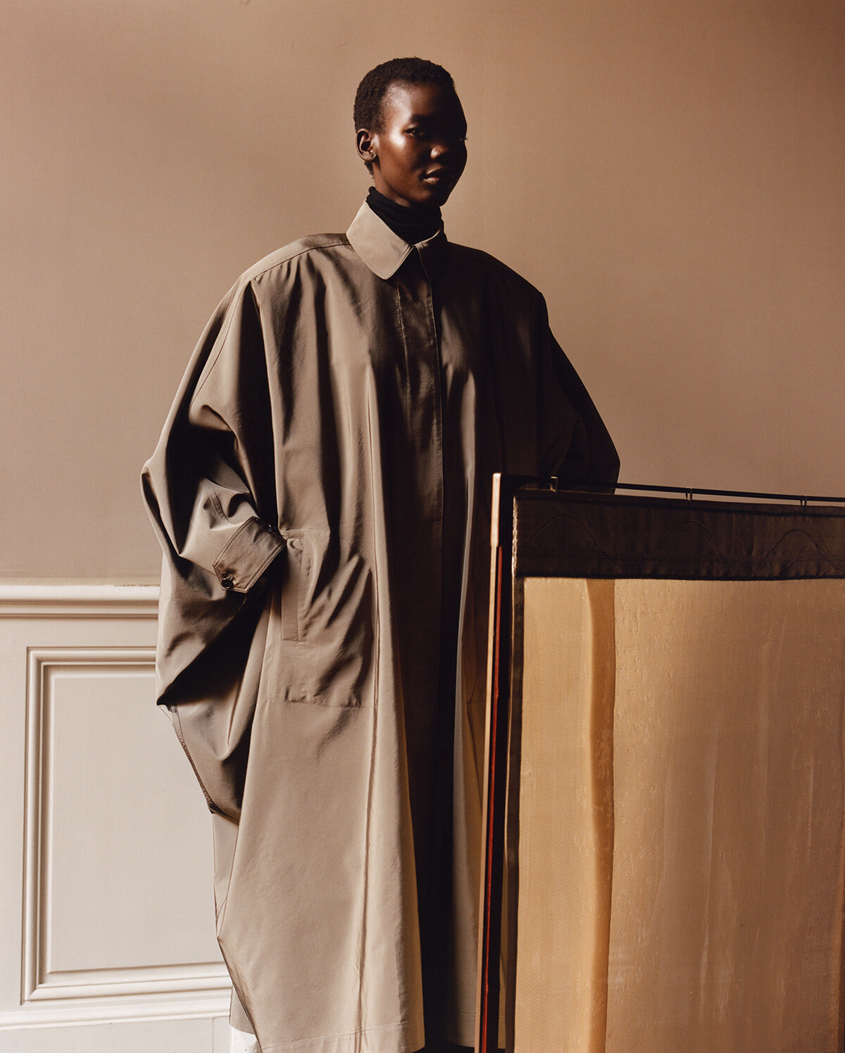 Person in an oversized beige trench coat standing next to a large folding screen in a softly lit room.