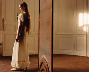Woman in a long cream dress with feathered sleeves stands in a sunlit room with white paneled walls.