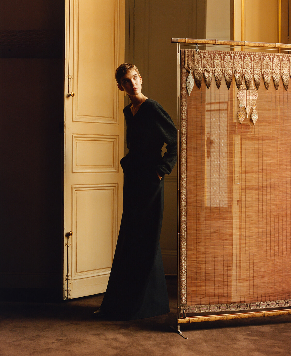 Woman in a long black dress leans from behind a pale doorway, gazing to the side in a warm, studio-like setting with a decorative bamboo screen nearby.