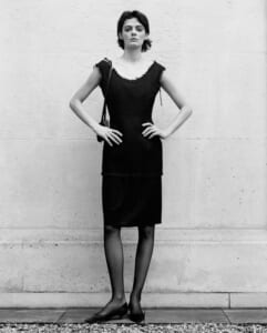Woman standing against a textured wall in a sleeveless black dress with a small shoulder bag, hands on hips.