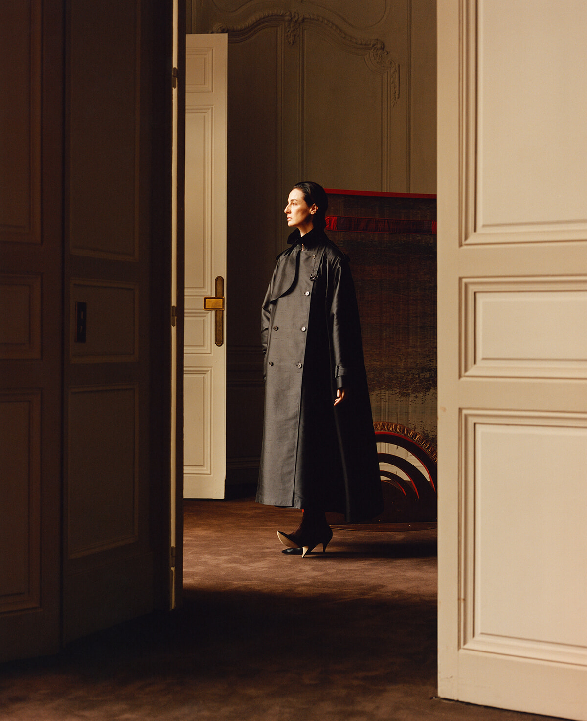 Woman in a long dark trench coat stands in a doorway of a richly paneled room, looking to the side.