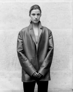 Person wearing an oversized coat standing against a concrete wall, hands clasped in front.