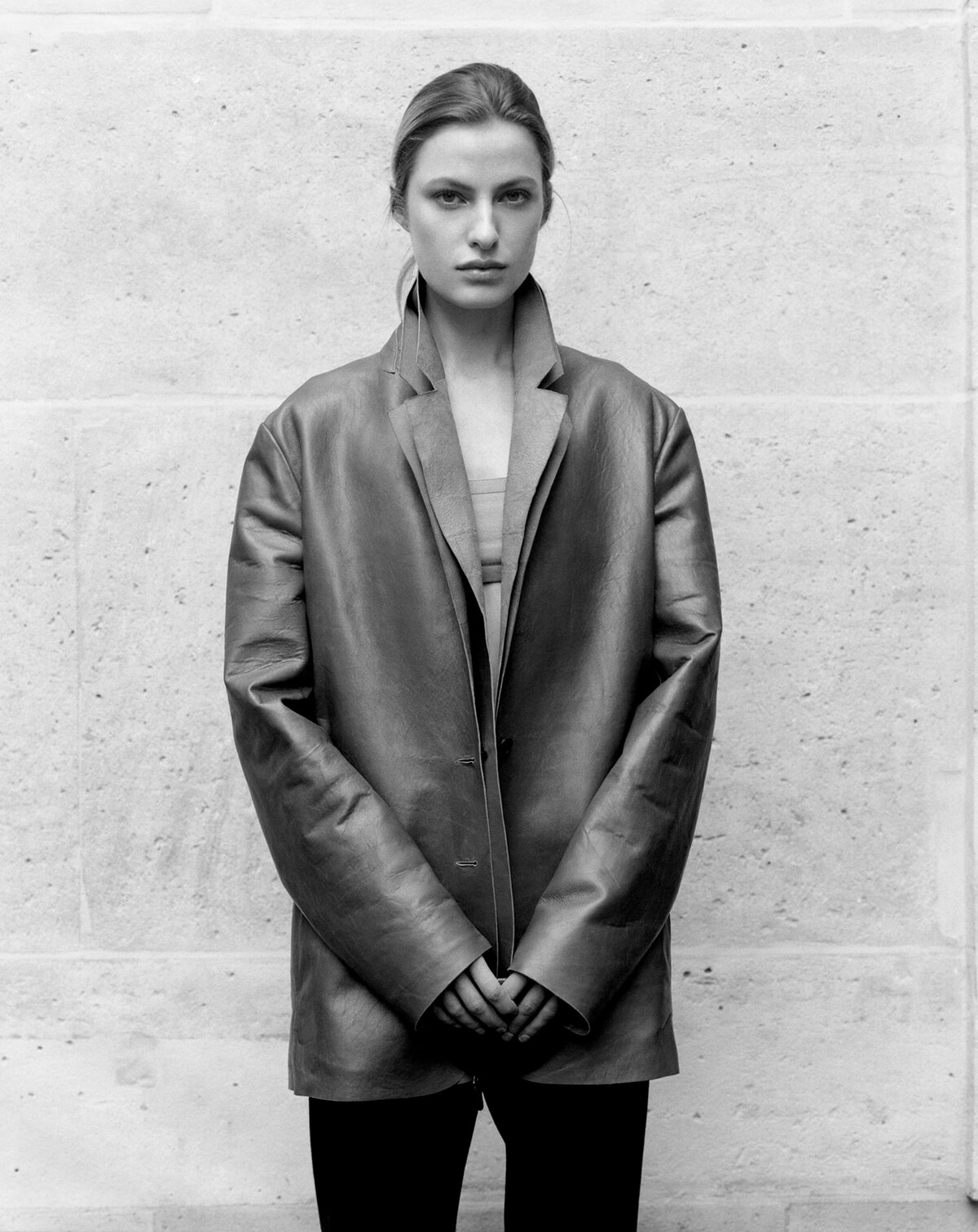 Person wearing an oversized coat standing against a concrete wall, hands clasped in front.