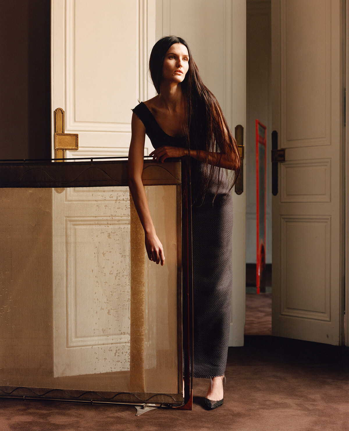 Woman with long dark hair leans on a wooden barrier in a stately doorway, wearing a black top and a gray patterned, full-length skirt, gazing off to the side.