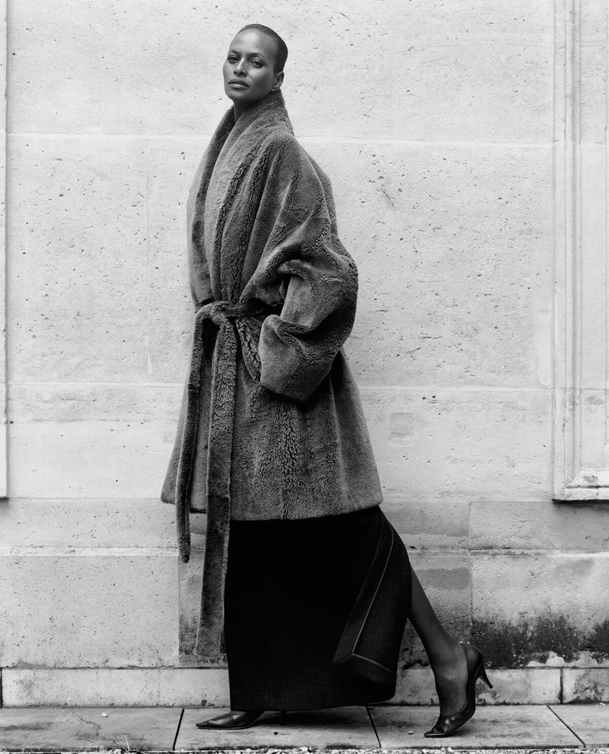Woman in a oversized fur coat with a belt, black pencil skirt, and heeled shoes walking along a stone wall; monochrome fashion photo.