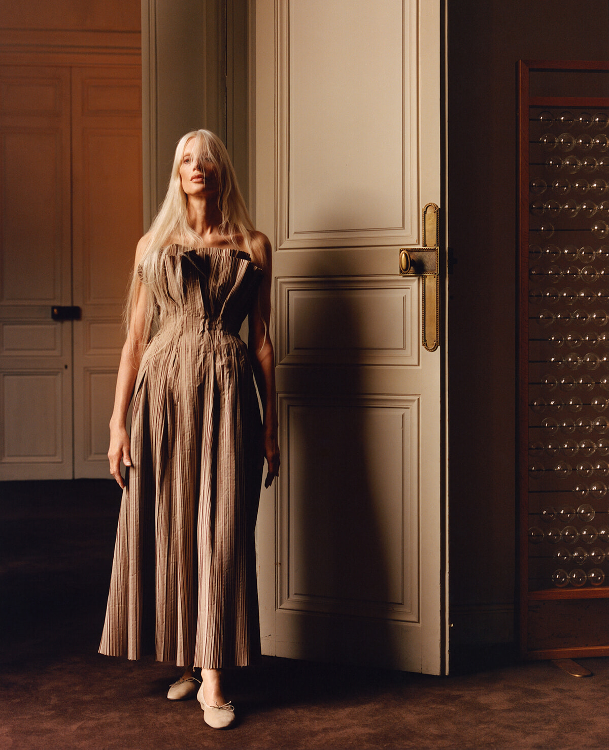 Woman in a beige striped, off-the-shoulder maxi dress stands by an open paneled door in a warmly lit room.
