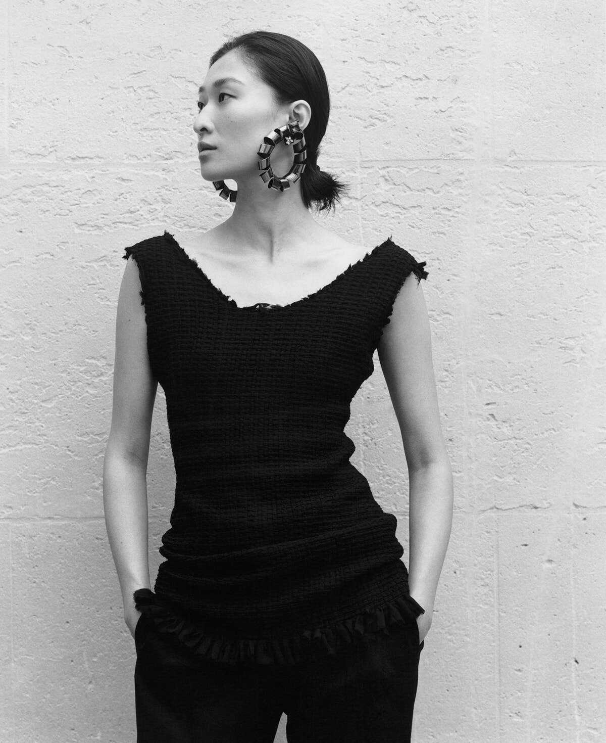 Woman wearing a textured black sleeveless top with oversized hoop earrings, standing against a light brick wall and looking to the left.