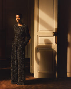 Person wearing a glittering black jumpsuit leans against a doorway, bathed in warm, golden light.