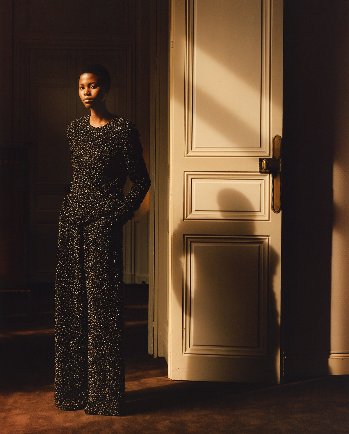 Person wearing a glittering black jumpsuit leans against a doorway, bathed in warm, golden light.